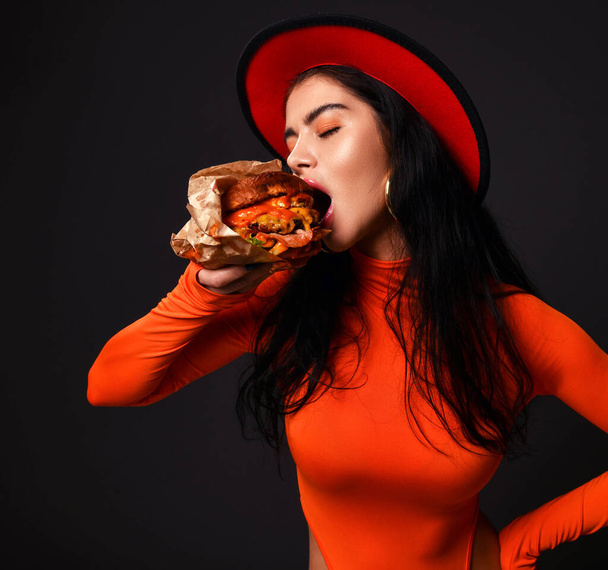 Голодна сексуальна пишна жінка з пишними губами в червоному тілі і капелюсі їсть кусаючи великий бургер з закритими очима
 - Фото, зображення