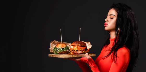 Jolie femme brune aux yeux fermés et aux lèvres boueuses en body rouge tient deux gros burgers frais sur un plateau en bois. Bannière - Photo, image