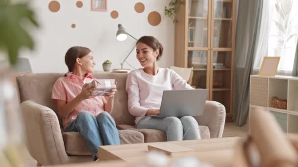 Uzamčení krásné smíšené-závod žena sedí na gauči v obývacím pokoji a pracuje na notebooku, zatímco její dospívající dcera přichází a dává jí dárek k narozeninám - Záběry, video