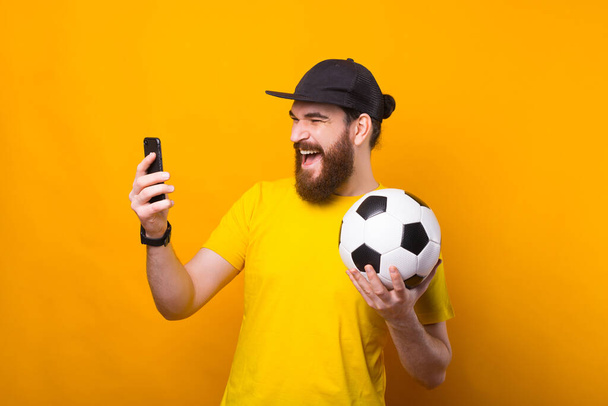 Portret zadziwiającego brodatego młodzieńca patrzącego na smartfona i trzymającego piłkę nożną, wspierającego ulubioną drużynę - Zdjęcie, obraz