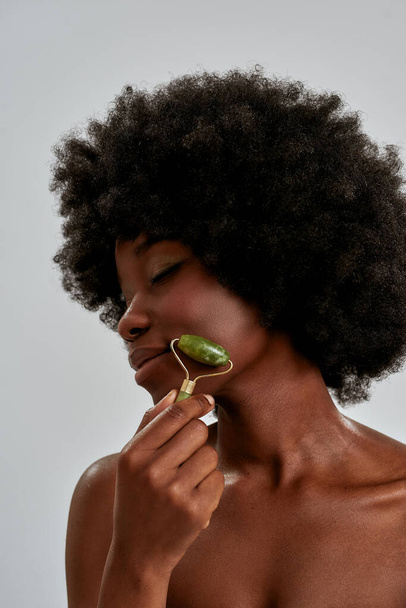 Retrato de cerca de una joven afroamericana relajada con una piel perfecta usando un rodillo de jade o una herramienta de masaje natural, posando aislada sobre un fondo gris - Foto, imagen