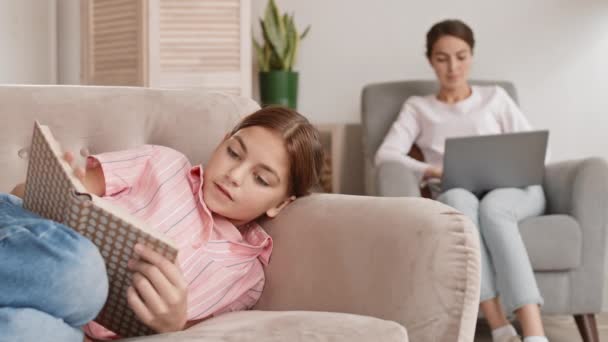 Κλείδωμα της έφηβης καυκάσιας κοπέλας ξαπλωμένη στον καναπέ στο σπίτι και διαβάζοντας ενδιαφέρον βιβλίο, ενώ η μαμά της κάθεται στην πολυθρόνα με το φορητό υπολογιστή στα γόνατά της στο παρασκήνιο - Πλάνα, βίντεο
