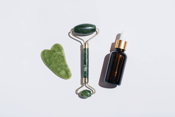 Jade face roller and glass bottle of beauty serum with shadows on on white background. Flat lay, top view. - Foto, imagen