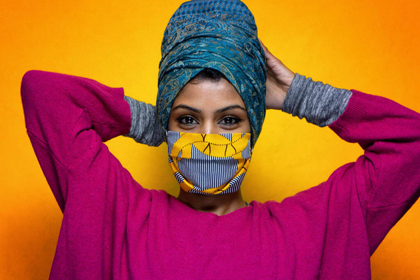 Beautiful young indian woman posing with covid 19 mask protection wearing a turban - Pretty Sri Lanka girl uses protection from pandemic flue posing with a yellow background - Photo, Image