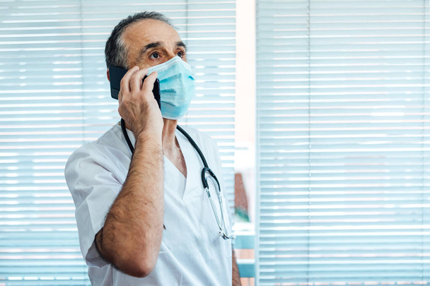 Reifer männlicher Arzt - Krankenschwester mit Gesichtsmaske, telefonierend neben einem Krankenhausfenster. Covid-19 und Medizinkonzept - Foto, Bild