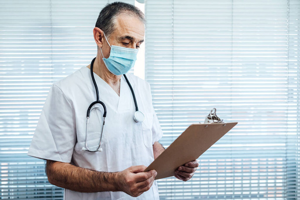 Зрелый врач-мужчина - медсестра в маске для лица, разговаривающая по мобильному телефону и держащая отчет у больничного окна. Covid-19 и концепция медицины - Фото, изображение