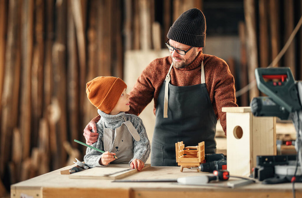 Hombre barbudo adulto en delantal y niño pequeño parado en el banco de trabajo y haciendo casa de pájaros de madera en taller de carpintería - Foto, Imagen