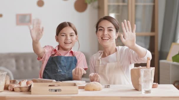 Mittlere Einstellung eines fröhlichen kaukasischen Teenagermädchens und ihrer gut aussehenden Mutter in Schürzen, die am Küchentisch sitzt, in die Kamera blickt, Mehl aus den Handflächen pustet und dann mit den Händen winkt - Filmmaterial, Video
