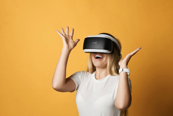 Excited happy screaming blonde woman in white t-shirt uses VR glasses exploring augmented reality holding hands up - Photo, Image