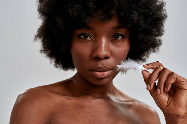 Close up portrait of charming african american woman with afro hair touching her facial skin with white feather, posing isolated over gray background - Фото, изображение