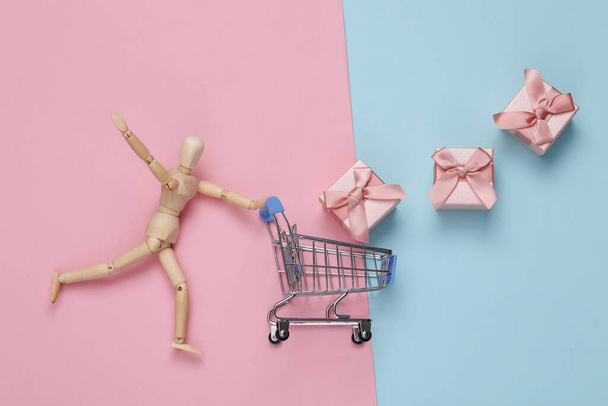 木製の人形はピンクブルーのパステルの背景にギフトボックスとスーパーマーケットトロリーを転がします。休日のショッピングコンセプト.  - 写真・画像