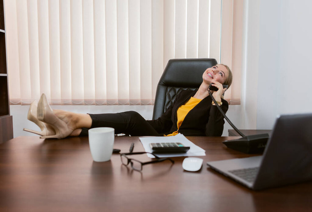 Розслаблена леді бос розмовляє по телефону і піднімає ноги на столі
 - Фото, зображення