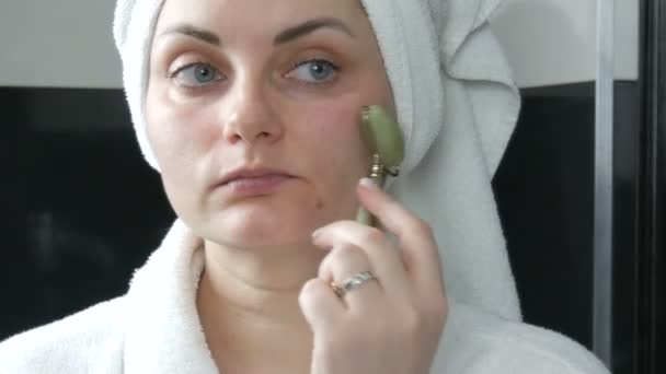 Krásná žena s ručníkem na hlavě při masáži s nefritovým válečkem na obličej přírodního zeleného kamene v koupelně. Čínské kosmetické nástroje. Lymfatická drenáž kůže masáž proti stárnutí vrásek - Záběry, video
