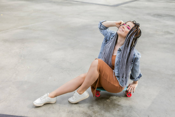 Extraordinary female millennial skater with colorful makeup, African braids sits on penny board and listen music in headphones at skate park. Youth, lifestyle concept - Photo, Image