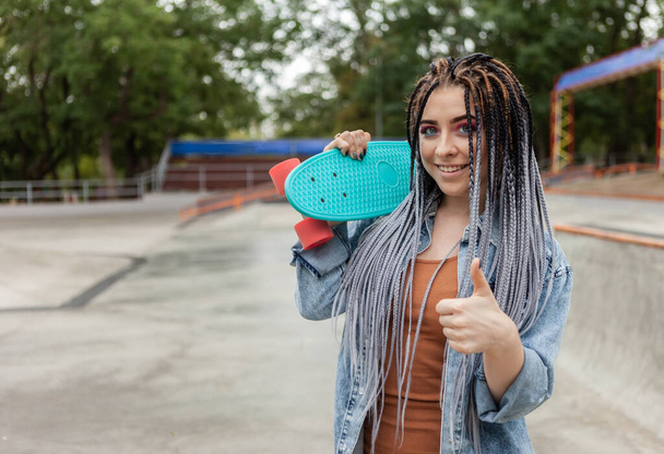 Fröhliche außergewöhnliche Millennial-Frau mit hellem Make-up und afrikanischen Zöpfen hält Penny Board in einem Skatepark - Foto, Bild