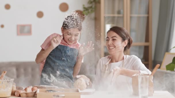 Medium shot of joyful Caucasian teenage girl and her good-looking mother wearing aprons sitting at kitchen table and having fun while making dough for a cake - Footage, Video