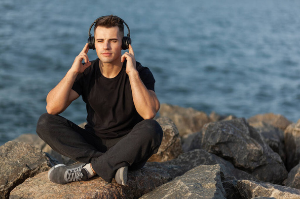 Healthy sport man listening to music while sitting on stones on the coast - Photo, Image