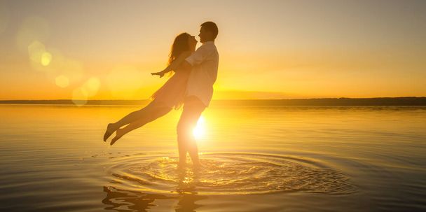 Young wedding couple is hugging in the water on summer beach. Beautiful sunset over the sea.Two silhouettes against the sun. Romantic love story. Man and woman in love in honeymoon trip. - Photo, Image