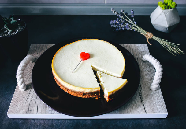 Classical homemade New York cheesecake is lying on dark-violet plate on wooden tray with rope handles on kitchen grey table. Lavender and succulent  on background. Red heart to Valentine's day present - Photo, Image