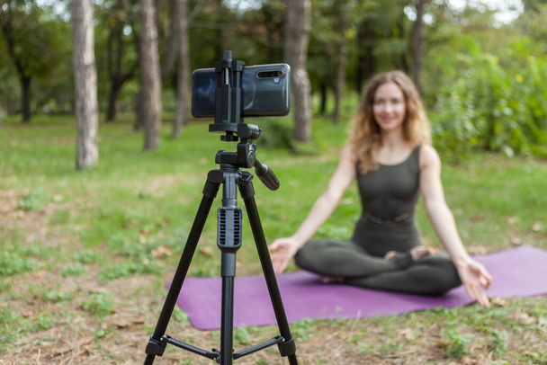 Giovane insegnante di yoga donna conduce lezione online nella foresta. La donna di sport trasmette a macchina fotografica di smartphone che sta su un treppiede. Blogging, vlog. Stile di vita sano - Foto, immagini