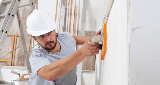 建設労働者は壁を滑らかにするためにスポンジとプラスチック製のトロールを使用しています。家の改善、内部の建設家のサイトで仕事で手の石膏の男とヘルメットを着用し、閉じる - 写真・画像