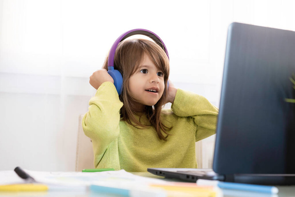Niña de la escuela primaria en auriculares sentados en el escritorio en el ordenador portátil - Sonriendo chica linda estudiando en línea desde casa y aprendiendo usando lecciones de Internet durante el brote de coronavirus - Concepto de educación en el hogar - Foto, imagen