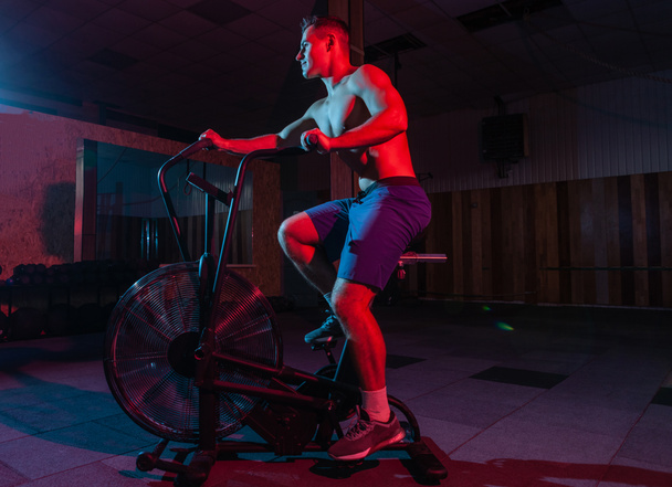 Entrenamiento de hombre muscular en bicicleta neumática en luz de neón rojo-azul. Formación cruzada - Foto, imagen