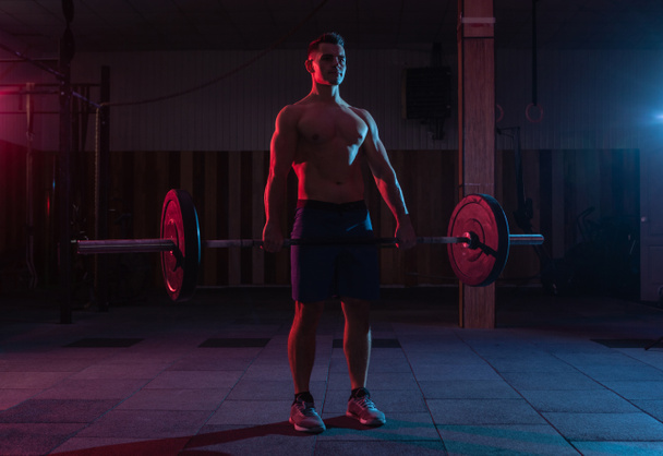 Тренируясь, поднимая тяжести в тренажерном зале. Сильный мускулистый человек делает тягу с тяжёлой штангой в красно-синем неоновом свете. - Фото, изображение
