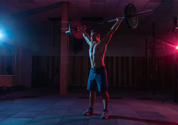 Muscular Caucasian man with muscular body doing barbell overhead press in a dark grungy gym with blue red neon light - Фото, зображення