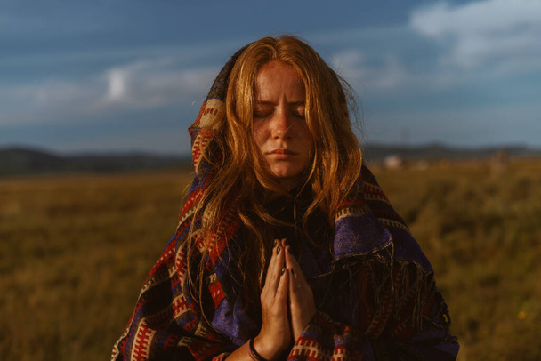 against the background of the low sky above the desert steppe with yellow grass, a girl in the cape of the eastern peoples prayerfully folded her eyes in meditation. She has red hair and freckles - Photo, Image