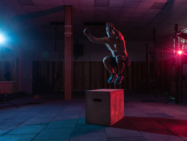 Ο μυώδης άνδρας προπονείται πηδώντας σε ένα ξύλινο κουτί σε ένα σκοτεινό γυμναστήριο. Αθλητής Γυμνάζεται σε κόκκινο μπλε νέον φως. Σταυρωτή, λειτουργική κατάρτιση - Φωτογραφία, εικόνα