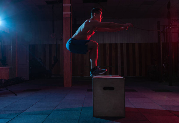 Homem musculoso treina pulando em uma caixa de madeira em um ginásio escuro. Atleta Treino em luz de néon azul vermelho. Cross, treinamento funcional - Foto, Imagem