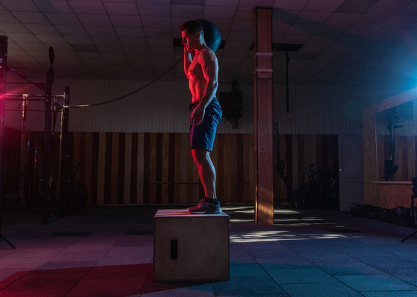 Svalnatý muž trénuje dřevěnou krabici přecházející s těžkou taškou v tmavé tělocvičně. Sportovní cvičení v červeném modrém neonovém světle. Zdravý životní styl křížení školení - Fotografie, Obrázek