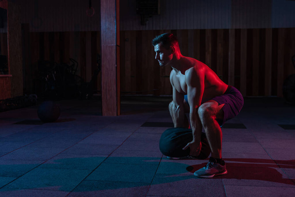 Ein muskulöser kräftiger Mann mit nacktem Oberkörper übt mit einer schweren Tasche im rot-blauen Neonlicht. Cross-Workout in einem modernen Fitnessstudio - Foto, Bild
