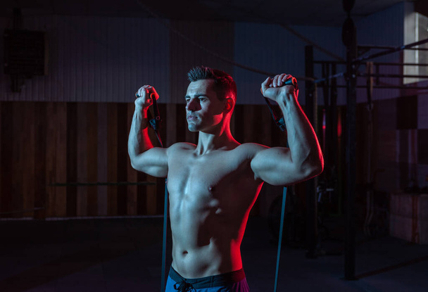 Hombre musculoso y poderoso con torso desnudo entrena con expansor (bandas elásticas de fitness) en luz de neón azul rojo. Entrenamiento cruzado en gimnasio moderno - Foto, imagen
