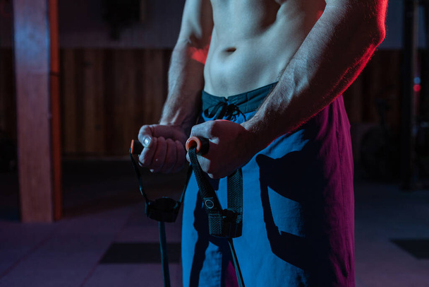 Lihaksikas voimakas mies, jolla on alaston vartalo, treenaa ekspanderilla (kuntokuminauhat) punaisella sinisellä neonvalolla. Cross harjoitus moderni kuntosali - Valokuva, kuva