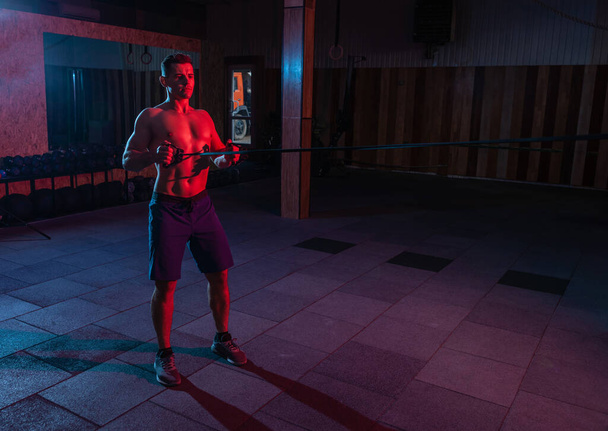 Spierkrachtige man met naakte romp traint met expander (fitness elastiekjes) in rood blauw neon licht. Cross workout in een moderne fitnessruimte - Foto, afbeelding