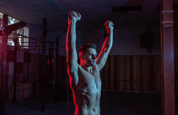 Muscular powerful man with naked torso trains with expander (fitness rubber bands) in red blue neon light. Cross workout in modern gym - Photo, Image