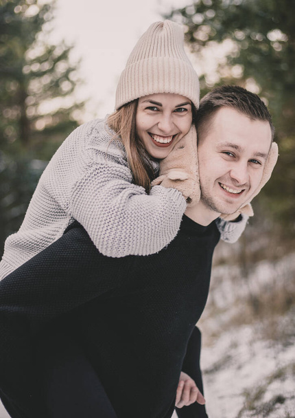 Μια γυναίκα με εμφάνιση μοντέλου κάθεται στην πλάτη ενός άντρα και αγκαλιάζει το λαιμό του στο φόντο ενός χιονισμένου δάσους. - Φωτογραφία, εικόνα