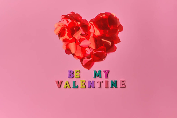 Be My Valentine lettering inscription for Valentine's Day greeting card. Red heart made of petals in shape of hearts, on pink background with inscription from multicolored wooden letters. Copy space - Photo, image