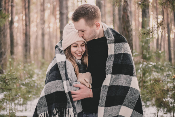 A man hugs and kisses a woman covered in a blanket against the background of a winter pine forest - Foto, Bild