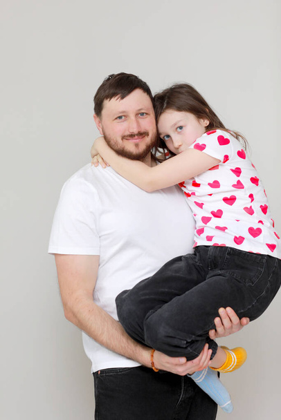 Nuori parrakas isä pitää tytärtään käsissään. Isä halaa lastaan rakkaudella. Mies valkoisessa t-paidassa ja tyttö punaisessa paidassa. Hyvää isänpäivää. Ystävänpäivä. - Valokuva, kuva