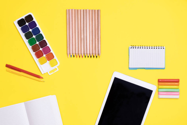 School background. Layout of educational supplies: tablet, alarm clock, paints, notebook, colored pencils on yellow background. Distance learning, children's creativity, online courses. Copy space. - Photo, Image
