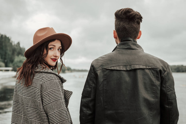 Back view of loving couple standing on the pier on lake or river in cloudy windy weather. Brunette lover and lady in hat spending time together. Image with copy space and blur background. - Фото, изображение