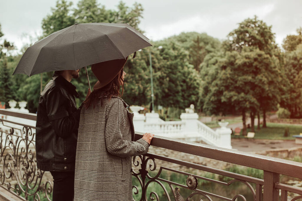Back view of heterosexual couple standing outside on the bridge in the city park near the river in cloudy rainy moody weather with black umbrella. Image with copy space and bokeh background. - Fotó, kép