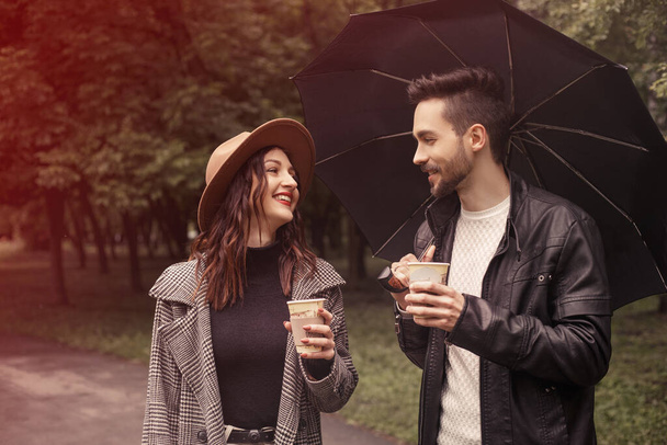 Νεαρό ζευγάρι περπάτημα άνοιξη πράσινο πάρκο της πόλης, πίνοντας ζεστό καφέ χαρτί οικολογικό ανακυκλώνουν κύπελλο. Δύο εραστές σύζυγος περνούν χρόνο μαζί, συζητούν ειδήσεις ταινία μαύρη ομπρέλα ταινία για να στεγάσει τη βροχή. - Φωτογραφία, εικόνα