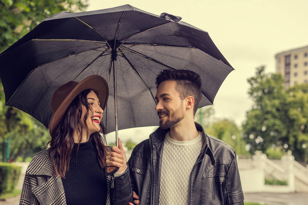 Portret młodej kochającej się pary spacerującej po parku miejskim i używającej parasola do schronienia przed deszczem. Heteroseksualni kochankowie mają randkę, spędzają razem czas w pochmurny deszczowy dzień, okazując emocje. - Zdjęcie, obraz