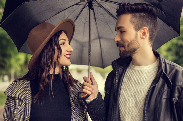 Retrato de una pareja joven y cariñosa caminando por el parque de la ciudad y usando un paraguas para protegerse de la lluvia. Amantes heterosexuales tienen una cita, pasar tiempo juntos en día lluvioso nublado, mostrando emociones. - Foto, Imagen