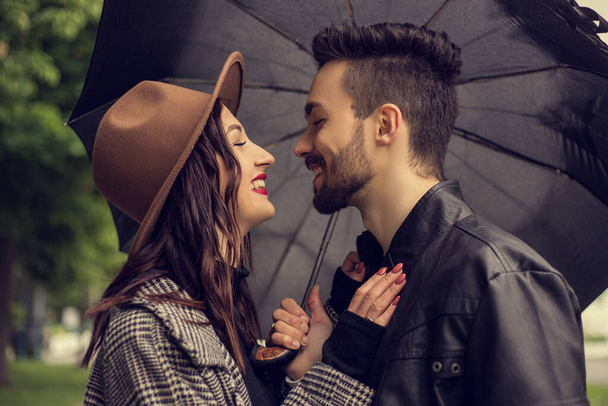 Portrait of young loving couple walking in the city park and use umbrella to shelter the rain. Heterosexual lovers have a date, spending time together in cloudy rainy day, showing emotions. - Photo, Image