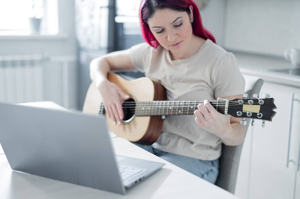 A woman sits in the kitchen during a remote acoustic guitar lesson. A girl learns to play the guitar and watches educational videos on a laptop - Фото, изображение